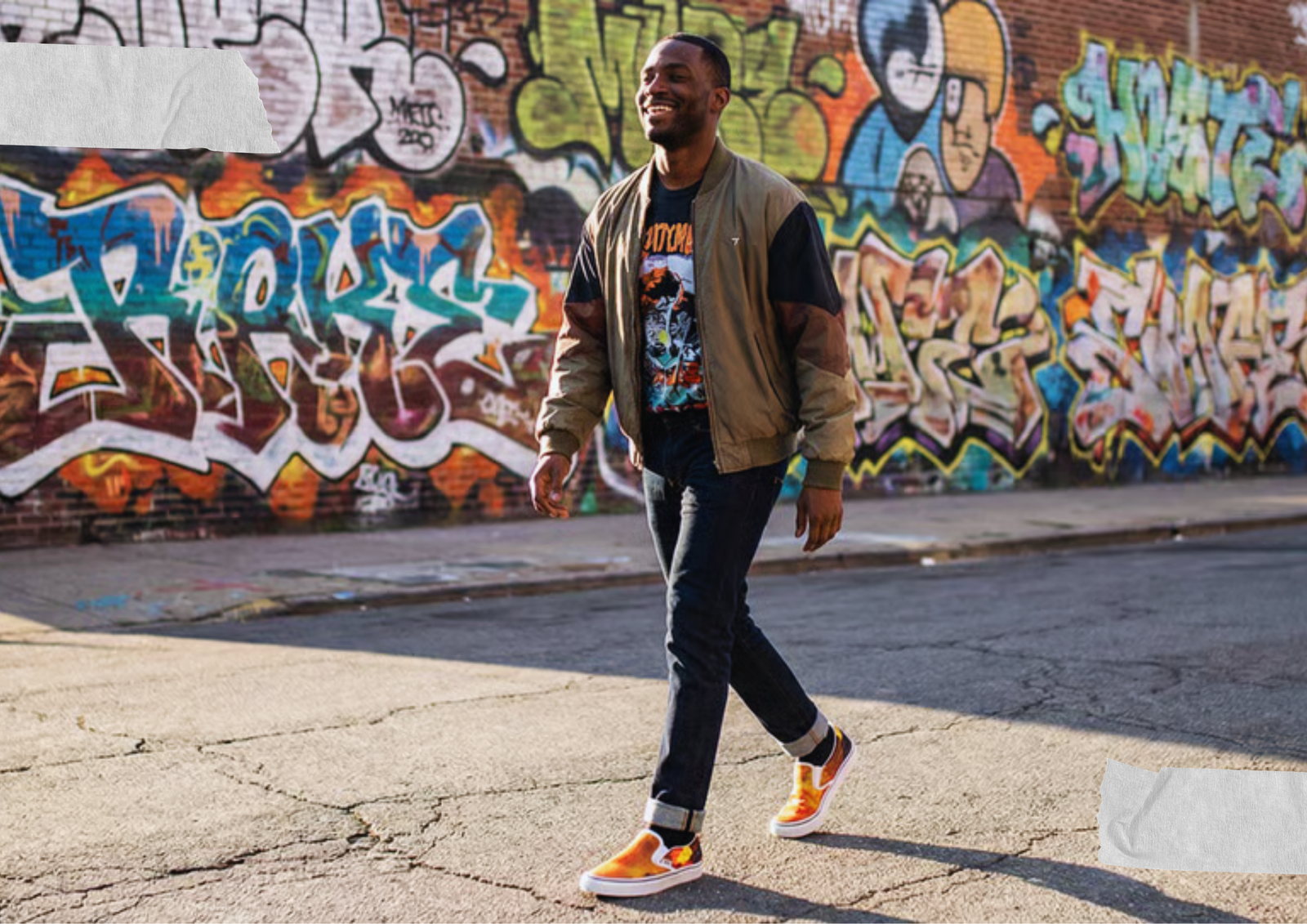 Man walking confidently down a city street wearing bomber girl slip-on sneakers, cuffed dark jeans, a black band tee, and a tan bomber jacket, with a colorful graffiti mural covering the brick wall behind him.