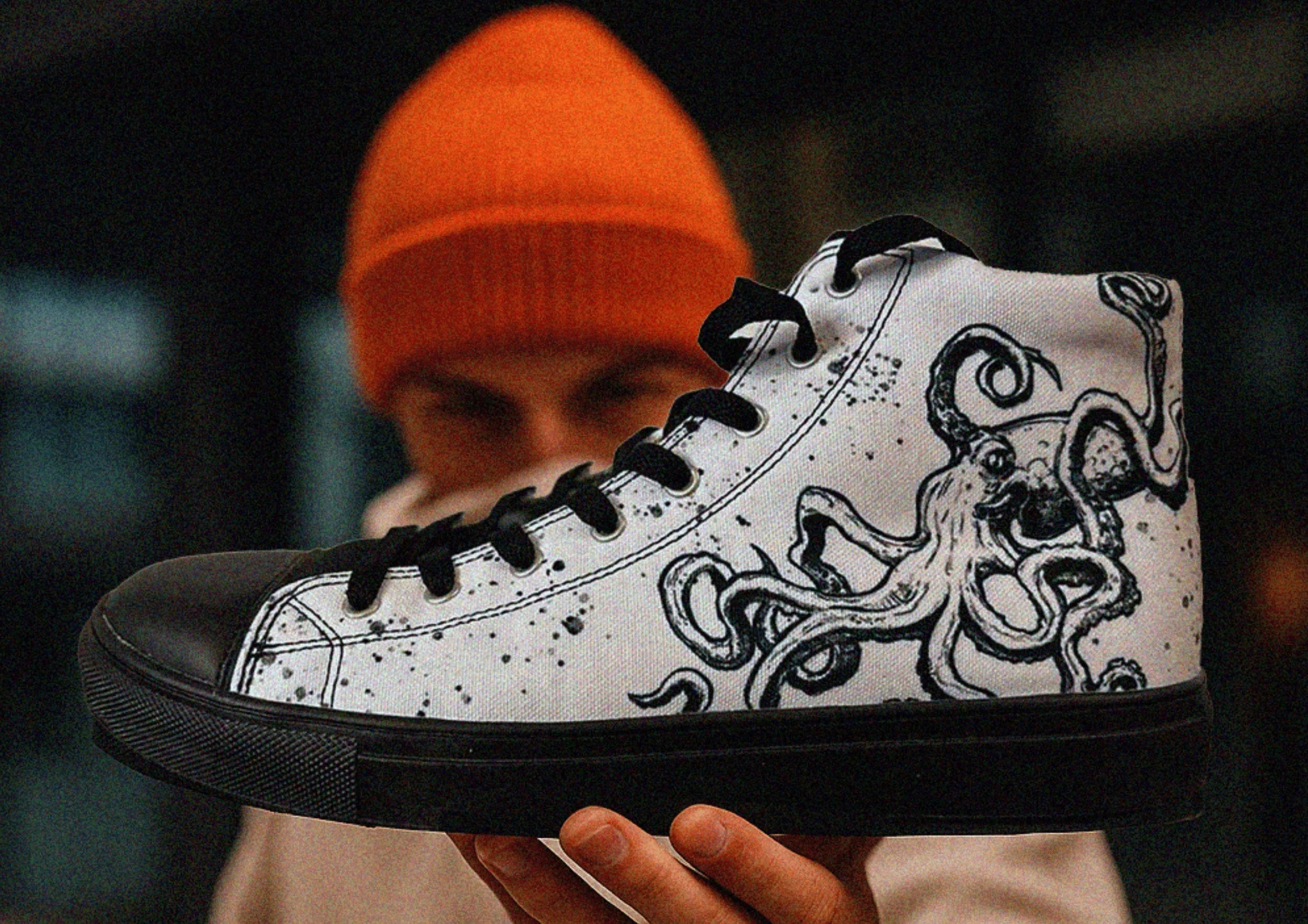 A man in an orange hat holding up a black and white high top canvas sneaker with an Octopus design by Sky Titan Media