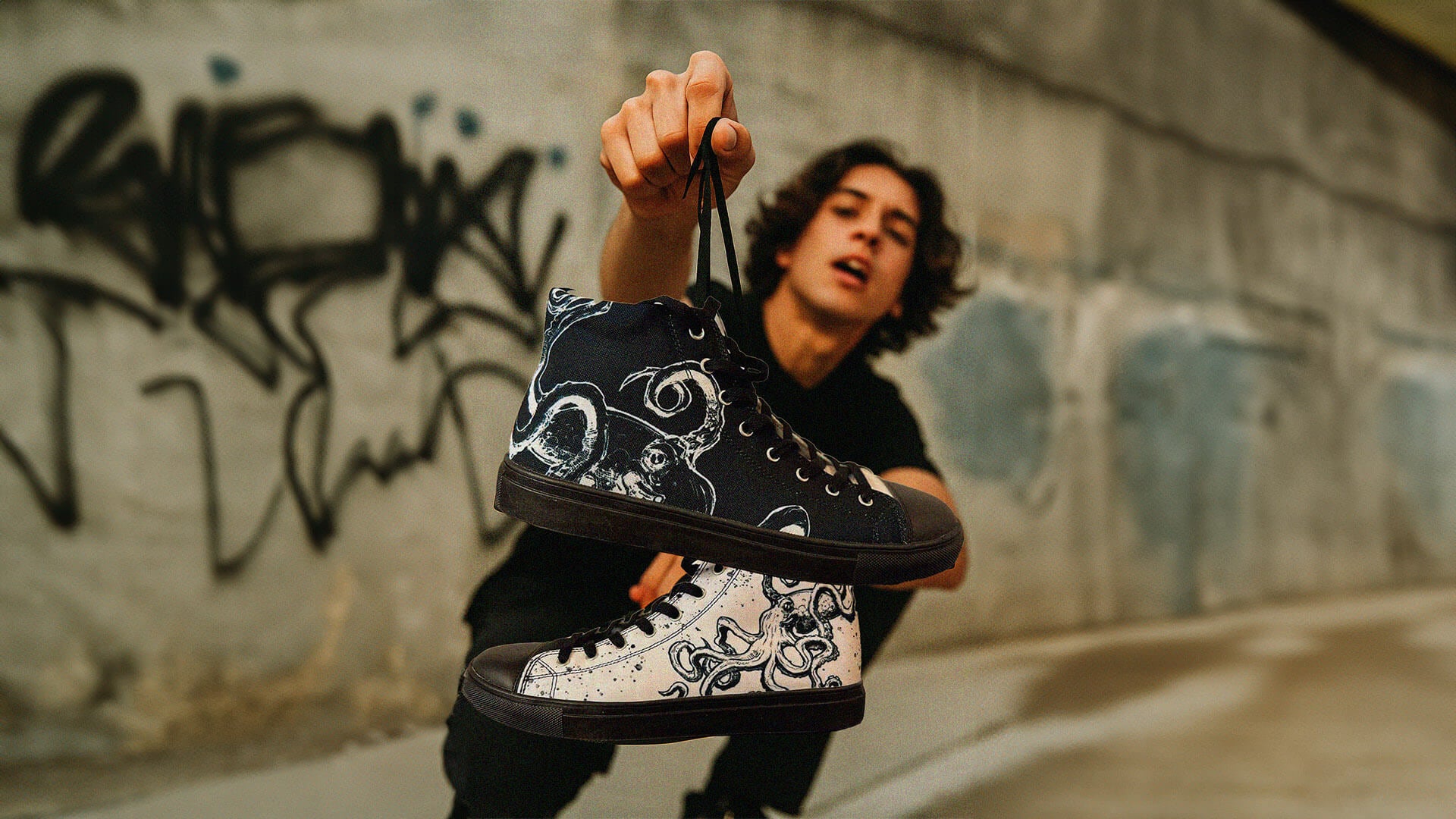 Person holding a black sneaker with white patterns against a graffiti-covered wall.