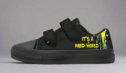 Kids’ black low-top sneakers featuring a vibrant neon cyberpunk-style cat illustration – Cyber Cat design by Sky Titan Media, built for young rebels with a love for futuristic flair and music-fueled streetwear.
Zoomed in, the left shoe makes its mark—“It’s A Mad World” jumps off the black canvas in a loud, neon yellow font. The black Velcro strap and toe box keep things grounded in style.