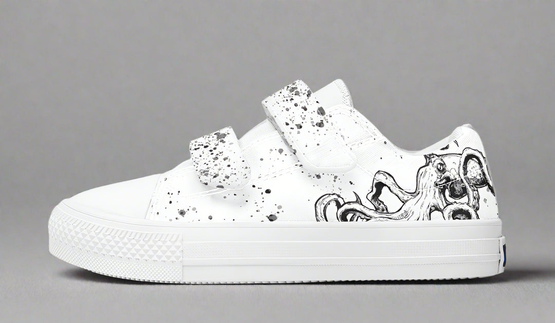 Side profile of the white-soled Octopus White Out Edition kids’ sneaker by Sky Titan Media, featuring hand-drawn black tentacle lines along the white canvas and a single bold velcro strap splashed with black ink. A stylish, kid-friendly silhouette that balances artistic expression with easy all-day wear.