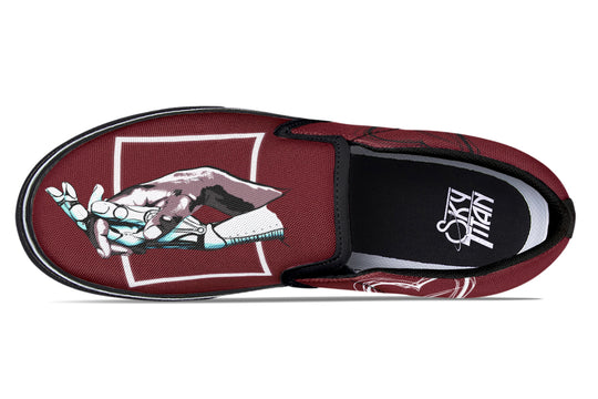 Overhead view of one red slip-on sneaker, displaying the full “July” design artwork—two robotic hands meeting within a sharp geometric box. The black elastic gore and white trim accentuate the central artwork.